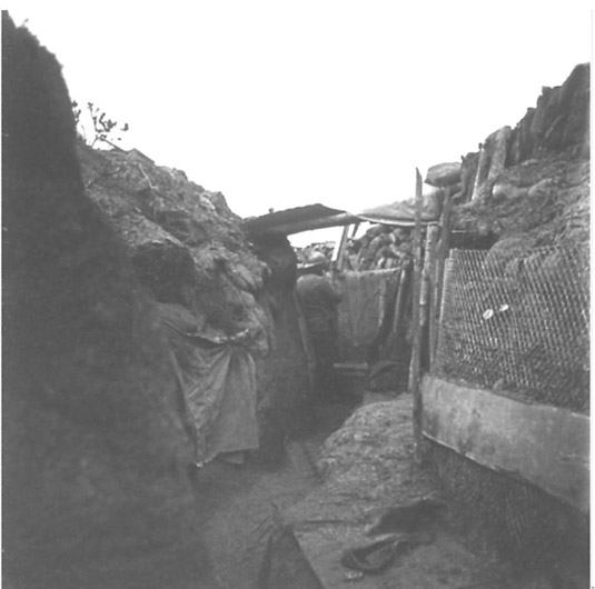 This photo displays the hodgepodge of material used in revetting a trench--planks, sandbags, chicken-wire, wattling. A small shelter is dug into the trench wall on the left with a tent cavas strung up as cover.