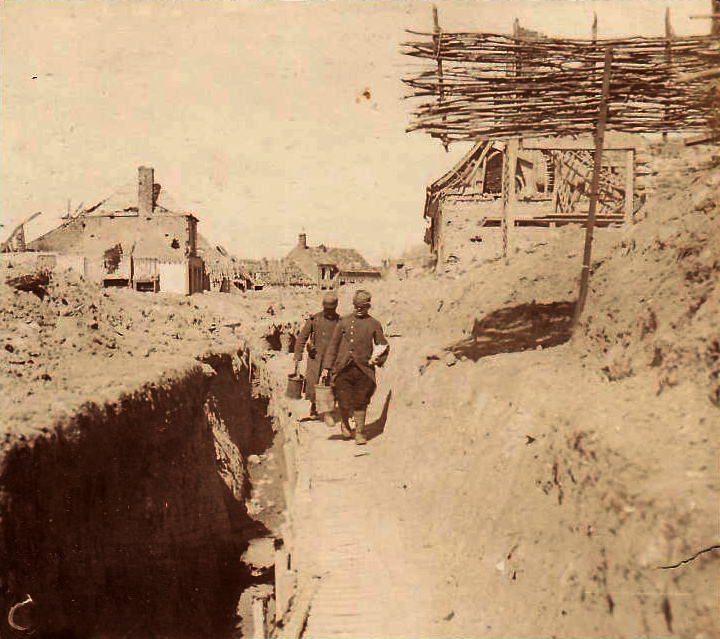 A ration party carries food along a trench that cuts through a town.